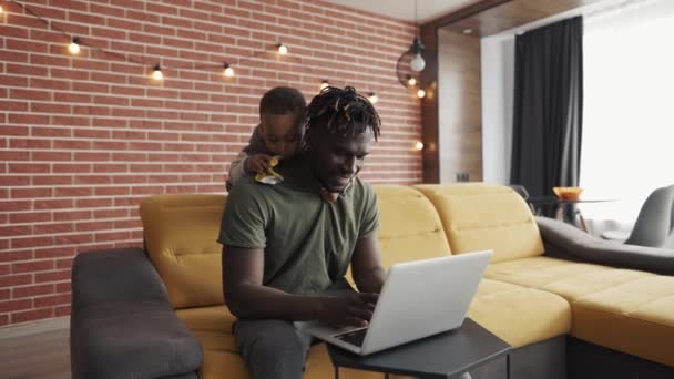Улыбающийся африканский отец занимается внештатной работой на ноутбуке, пока его сын беспокоит его сзади — стоковое видео