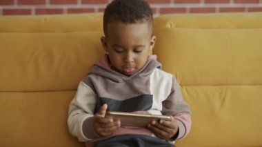 Afrikalı küçük şirin çocuk kanepede oturmuş akıllı telefon kullanıyor çizgi film izliyor ve eğleniyor.