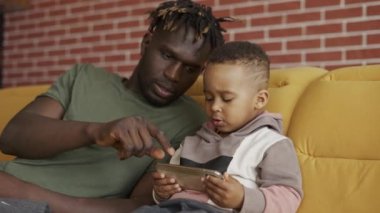 Afro-Amerikan babasının portresi. Oğluyla koltukta oturuyor. Akıllı telefon kullanan bir çocuk.