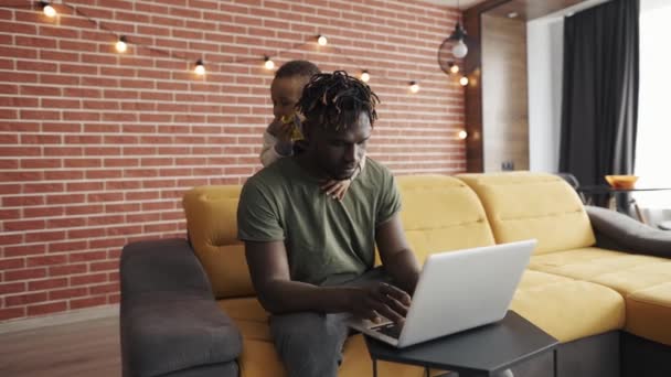 Drukke jonge Afrikaanse vader doet freelance werk op laptop terwijl zijn zoon hem van achteren stoort — Stockvideo