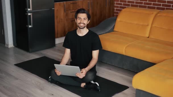 Portrait d'un homme assis sur le tapis sur le sol avec un ordinateur portable à la maison — Video