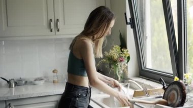 Parlak mutfakta akan suyun altında bulaşık yıkayan bir kadın.