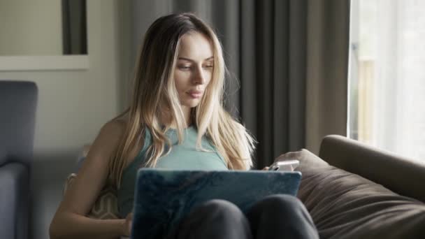 Blond vrouw zitten op de bank met laptop op de knieën met behulp van haar mobiele telefoon — Stockvideo