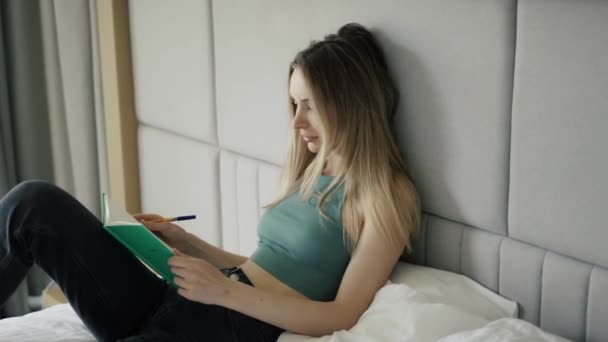 Mladá žena sedí doma na posteli a píše si do deníku — Stock video