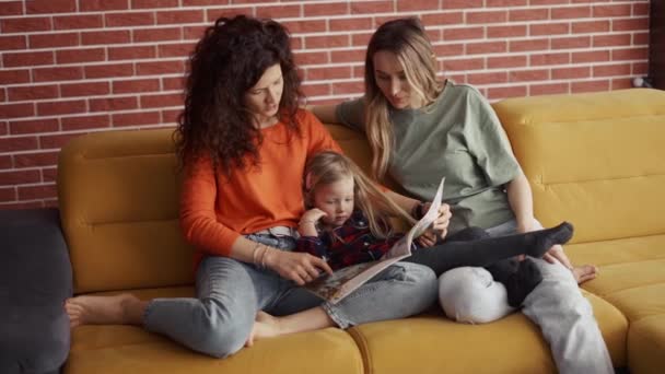 Les jeunes femmes lisent un livre ensemble à une petite fille d'âge préscolaire sur le canapé — Video