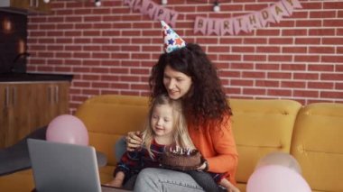 Anne doğum günü şapkalı anaokulu kızıyla oturuyor, bilgisayarından video görüşmesi yapıyor.