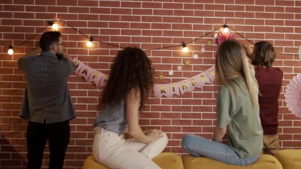 Amigos preparando fiesta en el interior colgando guirnaldas y cartas de cumpleaños, decorando — Vídeos de Stock