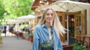 Ateşli sarışın kadın şehirde yürüyor, kameraya doğru dönüyor ve gülümsüyor.