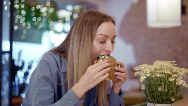 Frumos tanara blonda femeie mananca un burger mare într-un restaurant fast-food — Videoclip de stoc