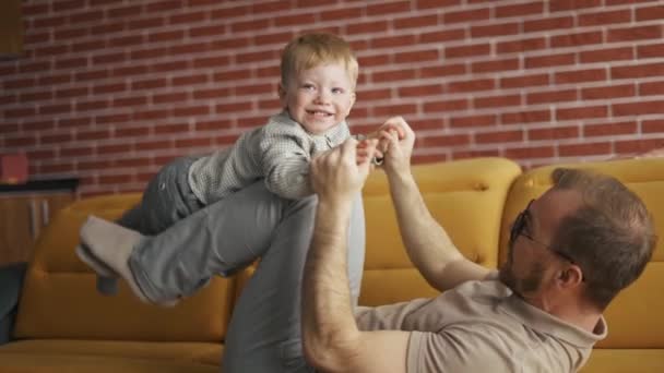 Hombre alegre sosteniendo saltando niño divirtiéndose juntos en el sofá en la sala de estar — Vídeo de stock