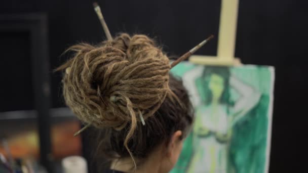 Veduta posteriore di pittrice con dreadlocks fissato da pennelli pittura su tela al cavalletto — Video Stock