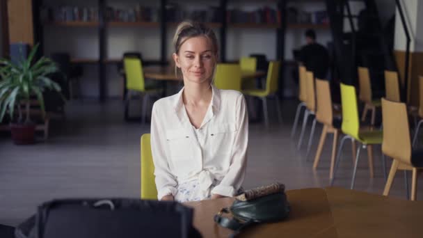 Porträtt av en ung kvinna leende, tittar på kameran mot bakgrund av ett bibliotek bokhylla — Stockvideo