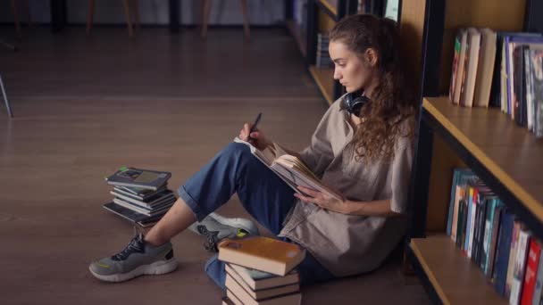 Estudante em fones de ouvido sentado contra estante com livros no chão — Vídeo de Stock