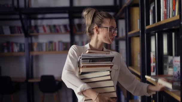 Blond kvinna håller hög med böcker i biblioteket, sätta mer på stacken — Stockvideo