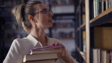 Çekici bir kadın kütüphanede kitap yığını tutuyor, başka bir kitap arıyor.