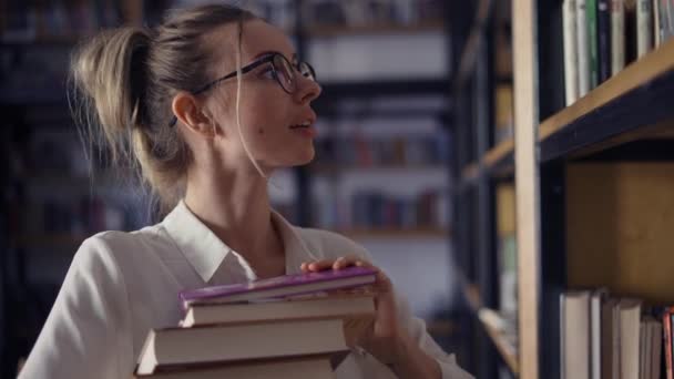 Mulher atraente segurando pilha de livros na biblioteca, à procura de outro — Vídeo de Stock