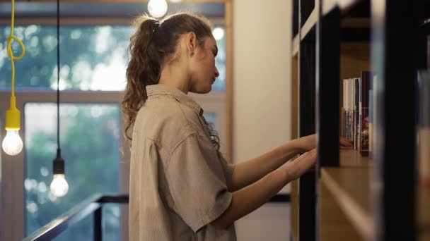 Student meisje kiezen van een boek in de bibliotheek op een boekenplank — Stockvideo