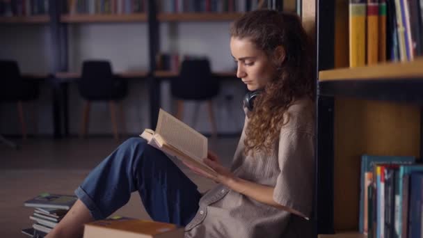 Student i hörlurar sittande mot bokhylla med böcker på golvet, närbild — Stockvideo