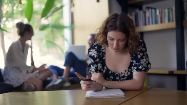 Mladá zamyšlená dívka sedí v knihovně a píše do sešitu — Stock video