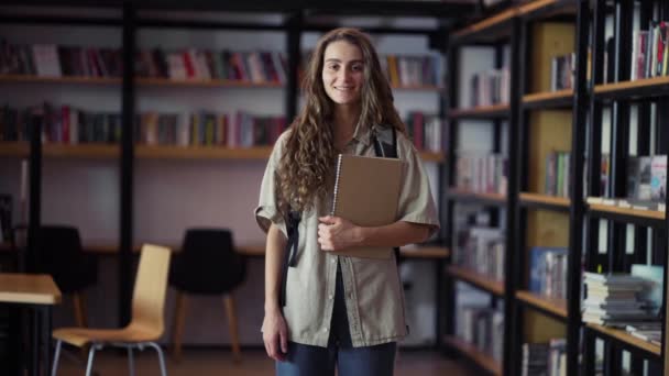 Portret van jonge mooie vrouw lachend gelukkig kijkend naar camera in bibliotheek boekenplank achtergrond — Stockvideo