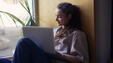 Kafede pencere kenarında oturan ve kulaklıkla dizüstü bilgisayarla çalışan serbest çalışan bir kadın.