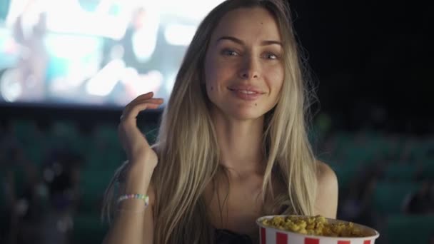 Donna bionda in piedi con secchio di popcorn al cinema, sorridente alla macchina fotografica — Video Stock