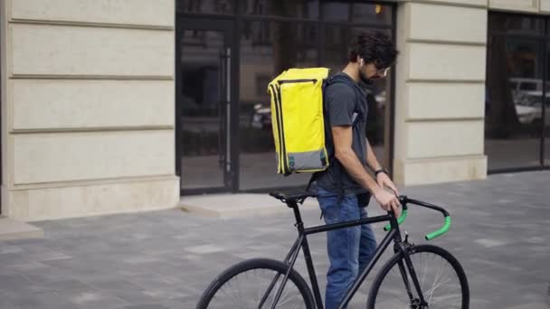 Bezorger met gele rugzak lopen op straat met een fiets, met behulp van oortelefoons antwoord op een oproep — Stockvideo