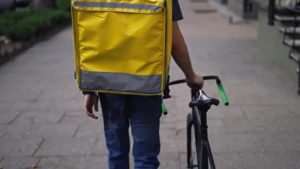 Entrega homem andando sozinho com bicicleta e saco amarelo, visão traseira — Vídeo de Stock