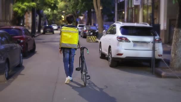 Liefermann geht allein mit Fahrrad und gelbem Sack durch die Abendstadt, Rückansicht — Stockvideo