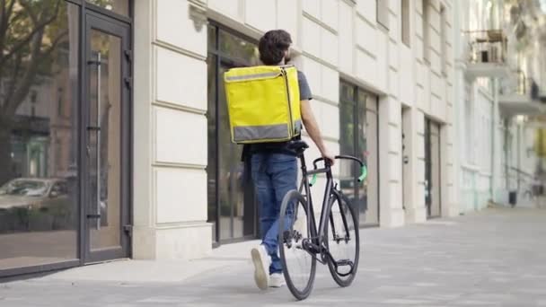 Entrega homem andando com bicicleta e saco amarelo, visão traseira — Vídeo de Stock