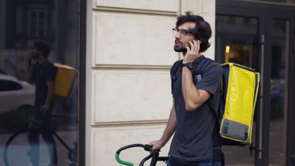 Курьер с желтым рюкзаком разговаривает с клиентом на улице, используя наушники, медлительно — стоковое видео