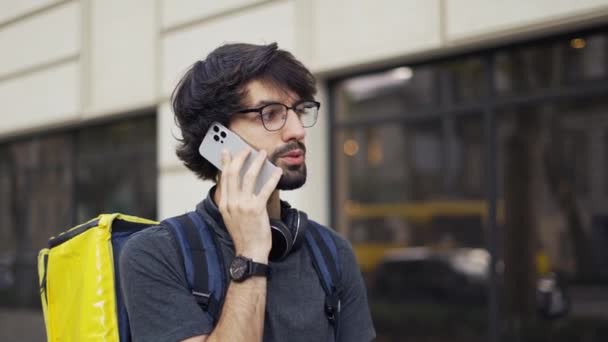 Liefermann mit gelbem Rucksack telefoniert mit einem Kunden auf der Straße — Stockvideo