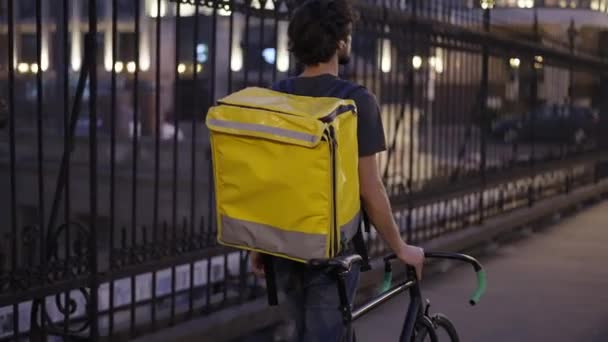 Entrega hombre caminando con bicicleta y bolsa amarilla por puente de la ciudad, vista trasera, cámara lenta — Vídeos de Stock