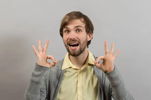 Está tudo bem! Jovem feliz na camisa gesticulando OK sinal e sorrindo enquanto está de pé contra fundo cinza — Fotografia de Stock