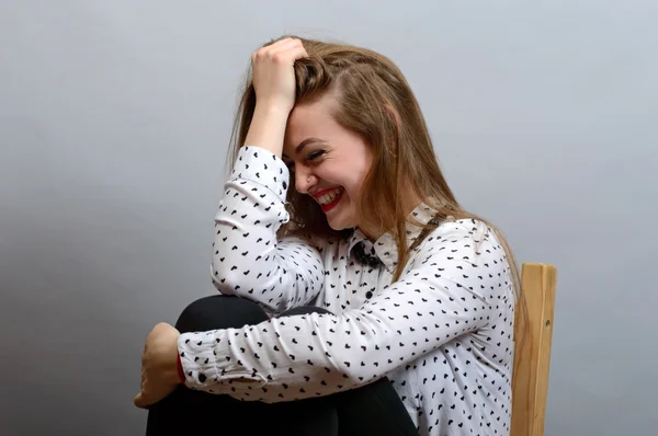 Hermosa mujer expresiva sentada en la silla y sonriendo sobre fondo gris . — Foto de Stock