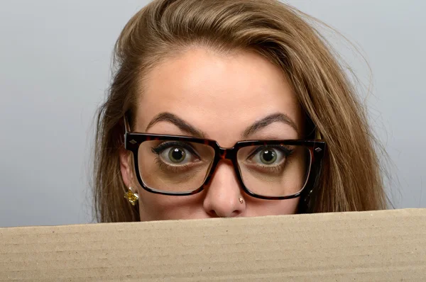 Eine Frau guckt über den Rand einer leeren Papierwerbetafel. schöne junge Frau mit Brille sieht überrascht und erschrocken komisch aus mit weit aufgerissenen Augen isoliert auf grauem Wandhintergrund — Stockfoto