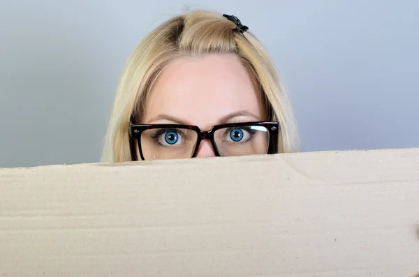 Banner sign woman peeking over edge of blank empty paper billboard. Beautiful young woman with glasses looking surprised and scared funny with wide open eyes isolated on grey wall background