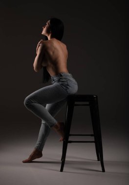 Düşünceli, üstsüz, esmer, uzun saçlı, bar sandalyesine yaslanmış bir kadın.