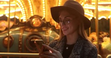 Şapkalı ve gözlüklü güzel bir genç kadın lunaparkta akıllı telefon kullanıyor. 