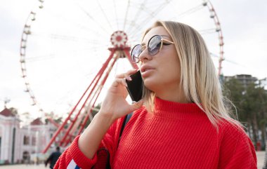 Kırmızı bluzlu ve güneş gözlüklü çekici sarışın kız lunaparkta akıllı telefon kullanıyor. 