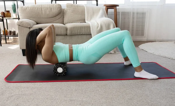 Fit sporty woman rolling on foam massage roller massaging muscles lying on yoga mat at home. Active african american girl doing fitness workout training exercise in living room