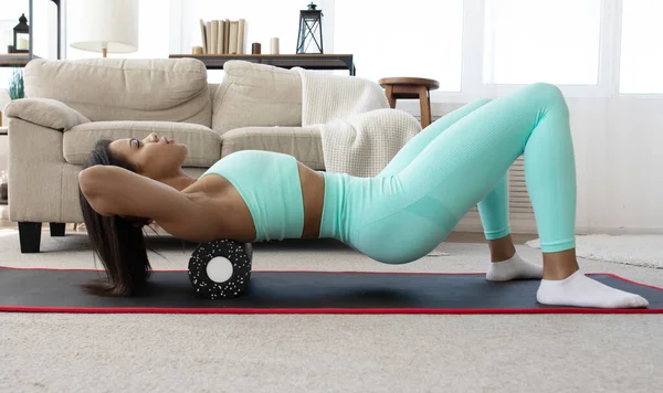 在家庭瑜伽垫地板上躺着穿着运动服在泡沫按压辊上滚动的健康的运动女性的侧视图 活跃的非洲裔美国女孩在客厅进行健身训练 — 图库照片
