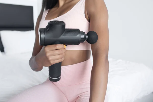 Evde perküsyon masaj aleti ile kaslara masaj yapan formda genç bir kadın.