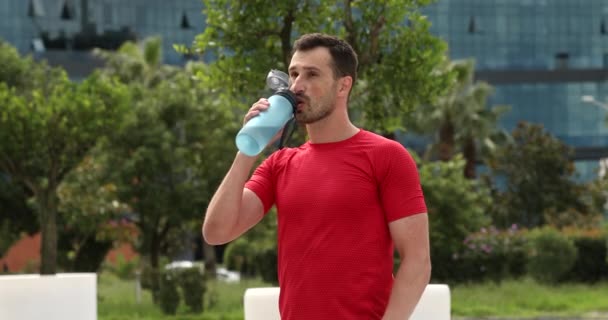身穿红色运动服的年轻肌肉男子在公园里行走 呼吸急促 喝着瓶子里的水 运动员在外面运动后 健康的概念 — 图库视频影像