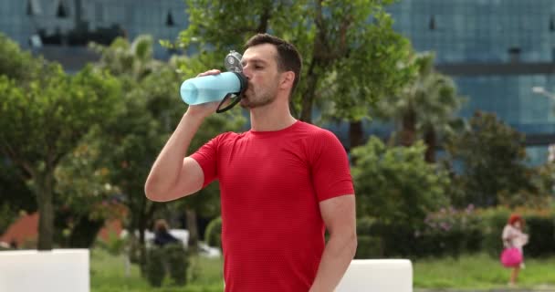 公園を歩いて呼吸する赤いスポーツウェアを着た若い筋肉質の男がボトルから水を飲んでいます 外でのトレーニングの後にアスリート フィットネスの概念 — ストック動画