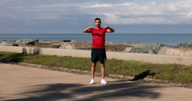Spor giyimli yakışıklı genç adam deniz kıyısındaki antrenmandan önce ısınıyor.