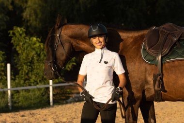 Gülümseyen genç kadın ata binmeye hazırlanıyor, eğitmen ata bakıyor..