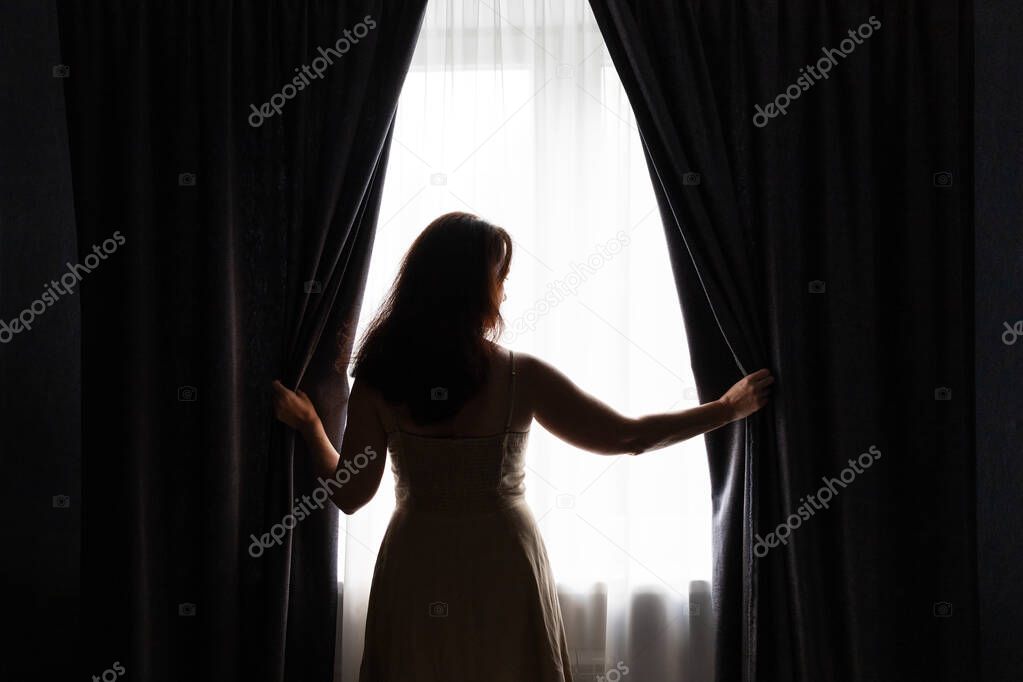A white woman is opening dark curtains and looking out of window, light coming to a room