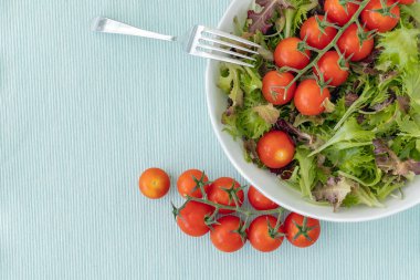 Karışık salata yaprakları ve kiraz domatesleri bir kasede ve açık mavi yüzeyde çatal, üst görünümde fotokopi alanı.