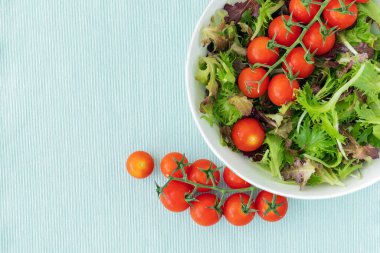 Açık mavi yüzeyde bir kasede karışık salata yaprakları ve kiraz domatesleri, fotokopi alanı ile üst manzara.